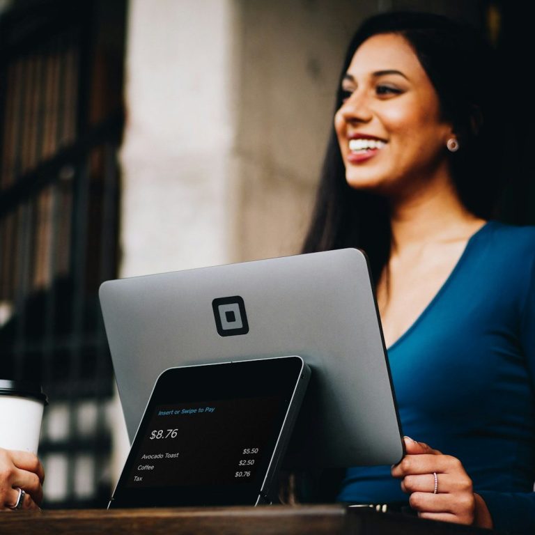 Smiling woman in a blue top, working on a laptop with a tablet and coffee nearby.