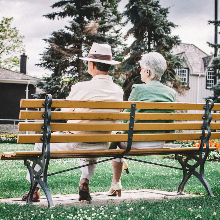 An elderly couple sits on a bench in a park, surrounded by trees and flowers.