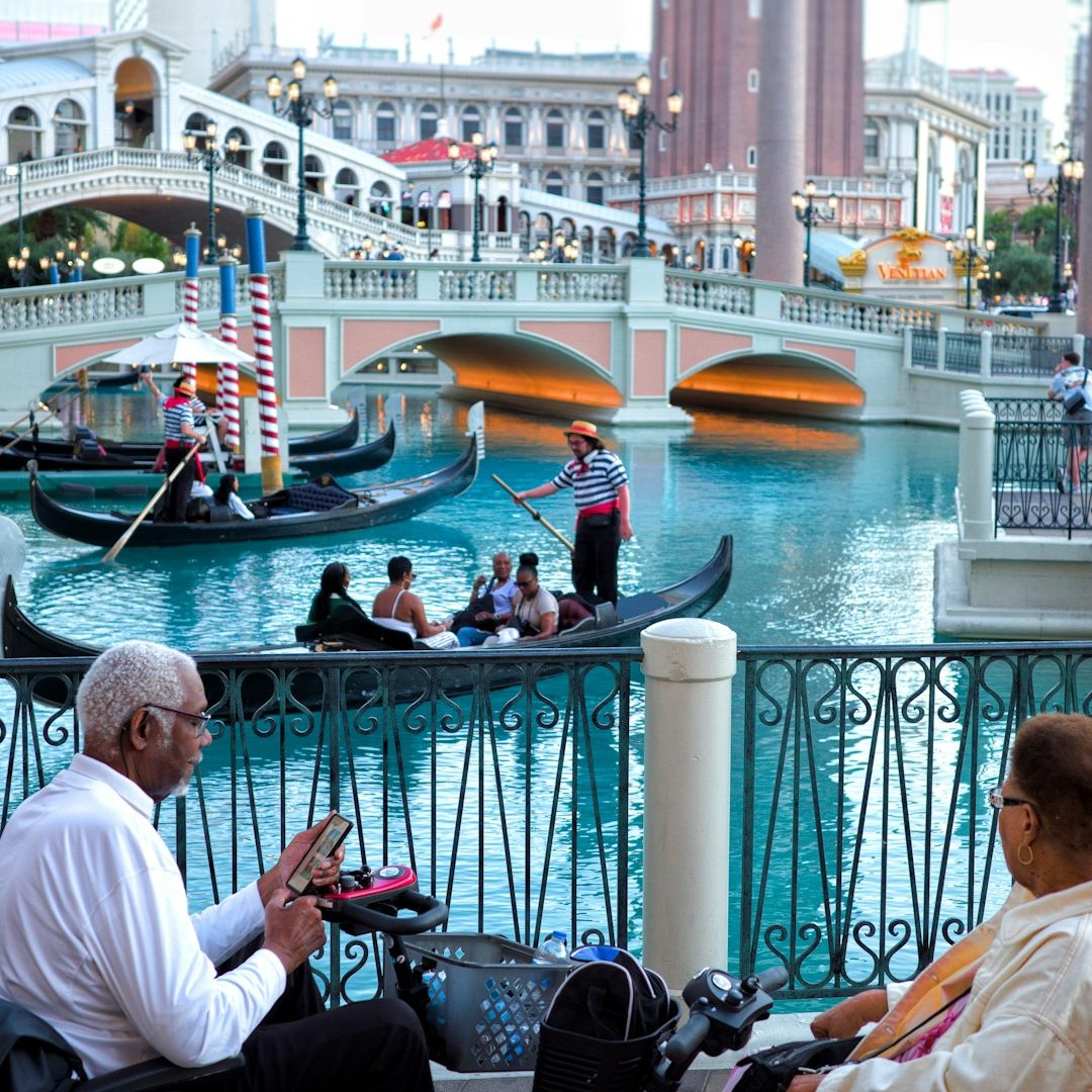 Gondolas on a canal, with people enjoying the scenery and a bridge in the background.