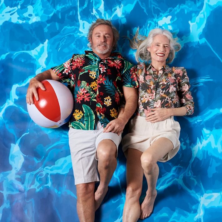 An older couple in floral shirts relaxing in a pool with a beach ball.