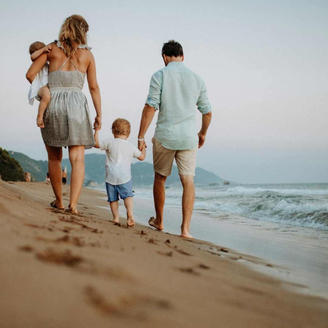 A family of four walking hand-in-hand along a sandy beach at sunset.
